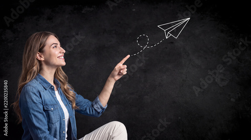 Smiling Young American Woman Pointing at White Paper Airplane Doodle on Dark Grunge Background