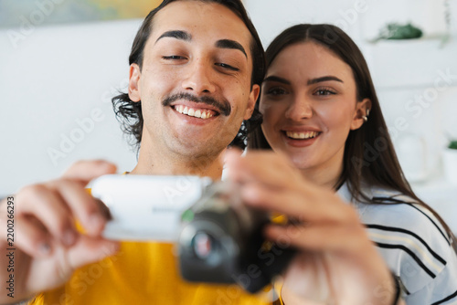 A smiling young man and woman stand together indoors while recording a video. They happily hold up a compact digital camera to capture a special moment at home.