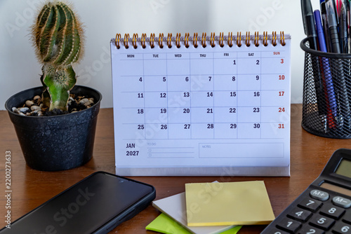 Desktop desk calendar for January 2027 surrounded by stationery, smartphone, cactus plant, sticky notes, and calculator.