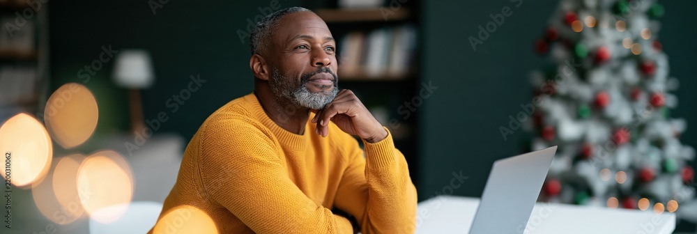 Obraz premium Pensive mature man working at home near a lit christmas tree with laptop for holiday banner and website background