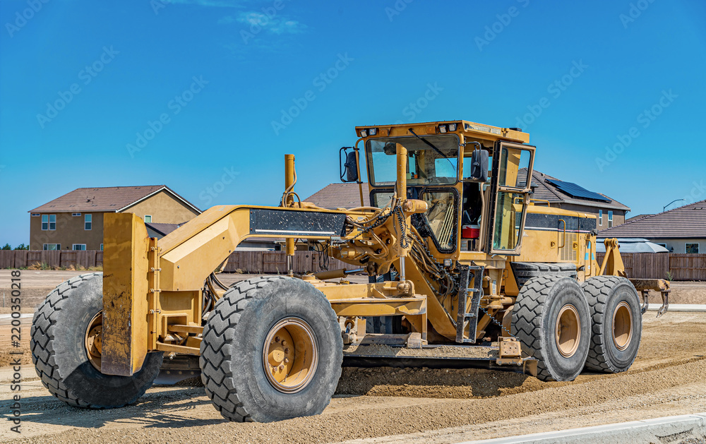 Fototapeta premium Heavy motor grader levels gravel for new roadway surrounded by residential construction. Land preparation.