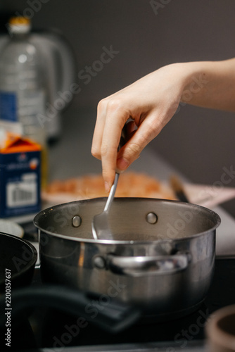 A close-up shot of a hand stirring food in a stainless steel pot on a stovetop, with a blurred background of kitchen ingredients and utensils, capturing a cozy home cooking moment