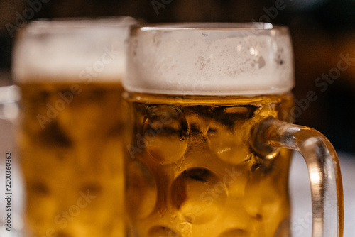 Close-up of beer mugs with thick foam, as a woman holds one glass and brings it to her lips. The image conveys relaxation, social drinking, and a cozy bar atmosphere.