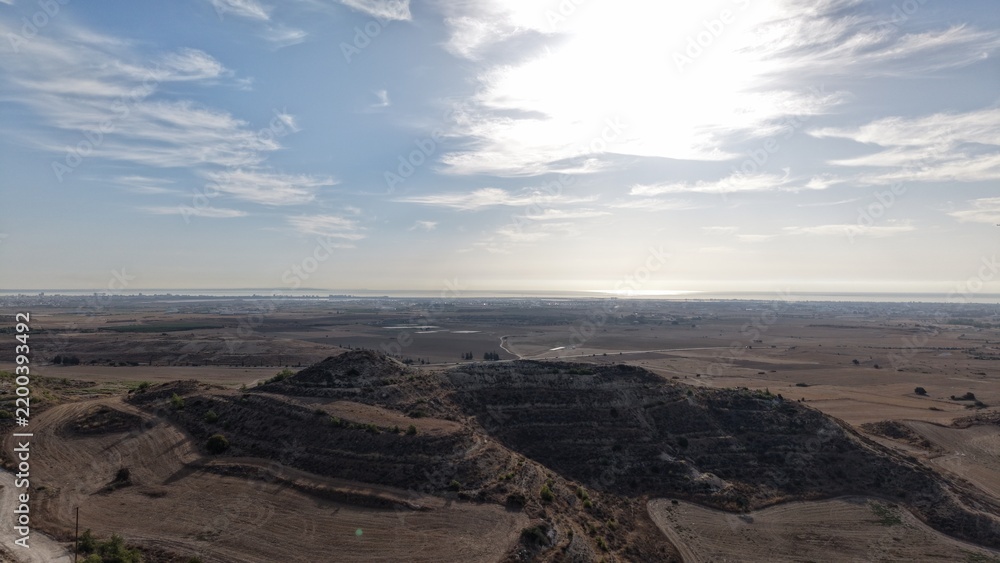 Fototapeta premium Aerial view of dry agricultural terrain in Cyprus with rocky hills, cultivated fields and distant coastal plains beneath a bright sky with scattered clouds
