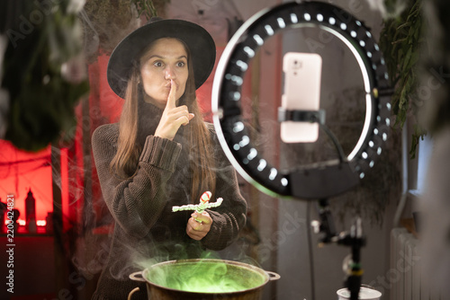 Halloween. witch blogger recording video with smartphone use cauldron prepare poison showing shh hush secret gesture. Black magic occult female wizard