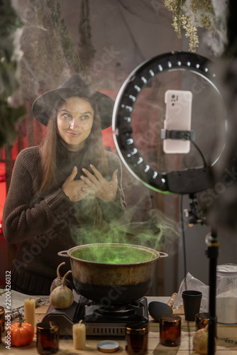 Halloween. Grateful witch blogger recording video with smartphone use cauldron prepare poison. Concept of empathy, gratitude, and emotional connection. Black magic occult female wizard