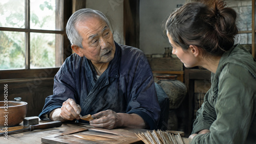 Elderly Japanese artisan demonstrating traditional woodcraft techniques to a young woman in a workshop