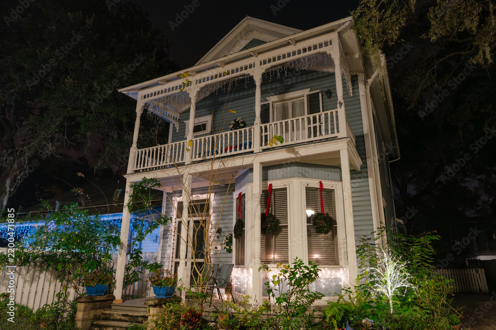 Fototapeta premium A blue Victorian house with white balconies and columns stands illuminated at night in St. Augustine, Florida. Christmas lights and wreaths decorate the windows, porch, and tropical garden.