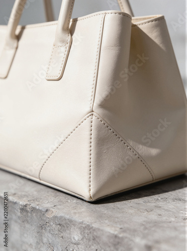 Cream leather handbag with precise stitching on a light gray concrete surface, close-up