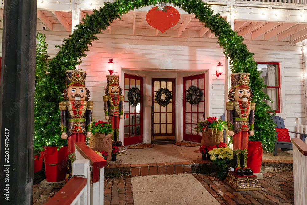 Obraz premium Three giant nutcracker soldiers stand beneath an illuminated garland arch in St. Augustine, Florida. Christmas trees, wreaths, poinsettias, and red ornaments decorate the historic building entrance.