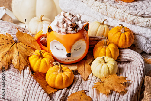 Creamy pumpkin spice latte topped with marshmallows and spices in Cute orange fox mug, surrounded by pumpkins and colorful autumn leaves.