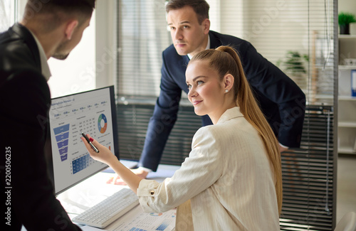 Woman analyst with colleagues reviewing data. She points to charts on a computer screen as colleagues plan strategy, review KPIs, and prepare a report. Concept is team analytics in office.