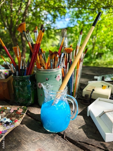 Outdoor artistic workplace setup with jars of various paintbrushes, a glass pitcher filled with blue water, paint palette, and miniature wooden houses on a sunny wooden table.