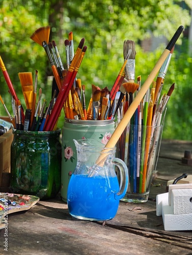 An assortment of creative painting tools and artist brushes placed in vintage containers next to a pitcher of blue water on a wooden workbench outdoors against a blurred green background.