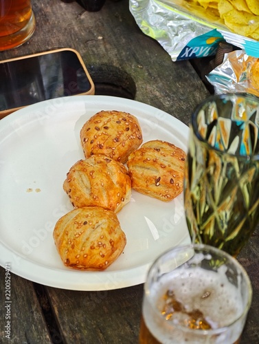 Golden baked puff pastry bites sprinkled with sesame seeds served on a white plate alongside cold beer, snacks, and a mobile phone on a wooden picnic table outdoors.