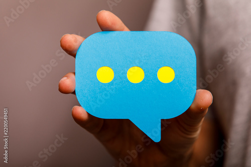 A person's hand holds a blue digital speech bubble with yellow dots, symbolizing online
