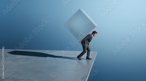 A businessman in a suit carrying a heavy large square cube on his back while walking on the edge of a platform