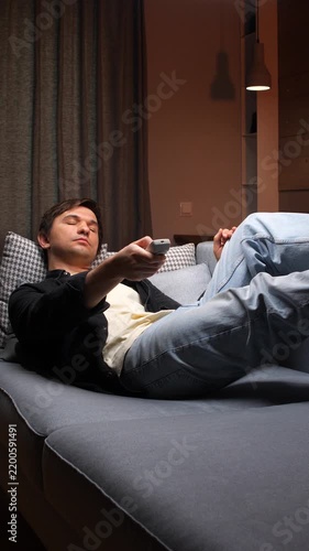 Vertical video young man lying on a comfortable sofa, holding a tv remote control, enjoying home entertainment and unwinding after a long day in his living room