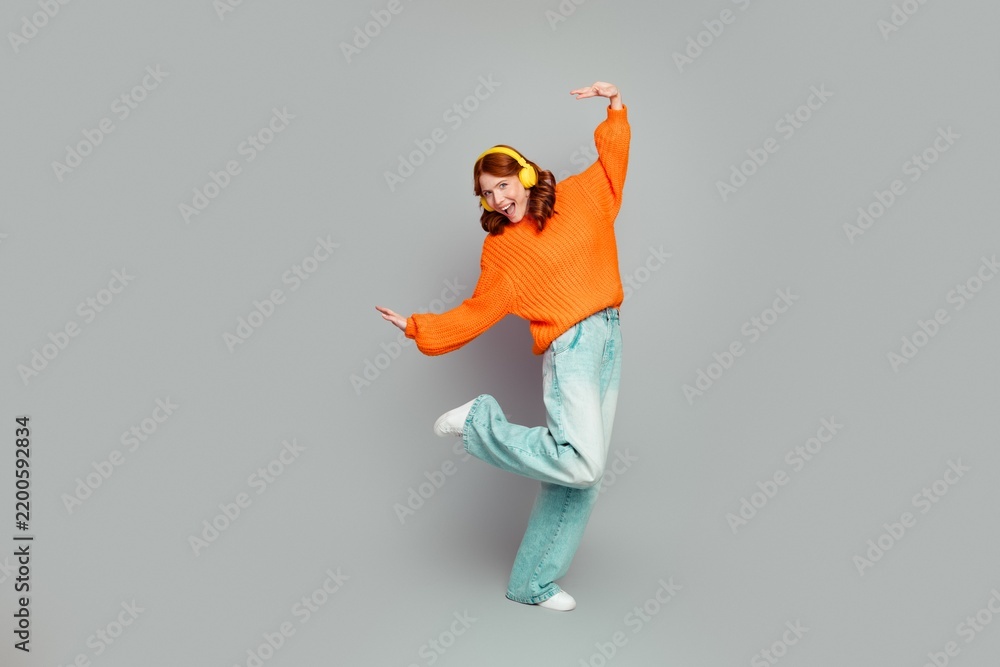 Obraz premium Young woman dancing in an orange sweater with yellow headphones in a photography studio, enjoying music, playful energy and casual denim streetwear