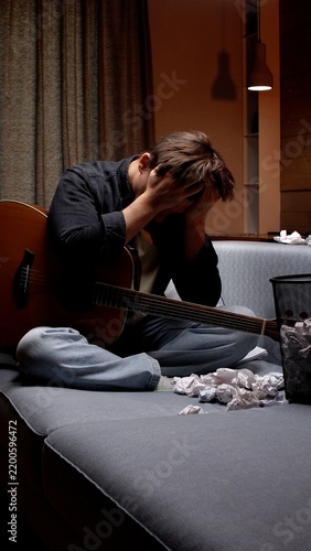 Young man sitting on a couch, holding his head in frustration next to a guitar and crumpled paper, feeling the exhaustion and stress of a creative block while songwriting