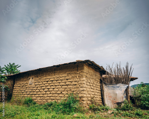 View of a traditional mud-brick house with a thatched roof and a makeshift fence made of branches and fabric in a rural landscape in Jos East, Plateau, Nigeria.