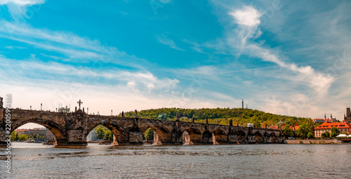 View of the historic Charles Bridge spanning the Vltava River with the city skyline in the background under a bright blue sky in Prague, Prague, Czechia.