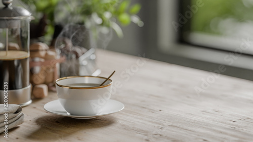 Steaming cup of coffee with gold rim on saucer beside French press on wooden table, blurred greenery and window in background.