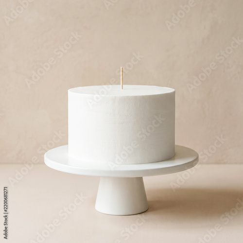 Minimal white frosted celebration cake on a white pedestal stand with a blank wooden topper stick