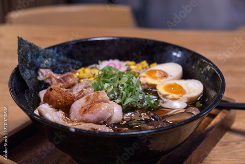 Bowl of ramen with sliced pork, soft-boiled eggs, green onions, and nori served on a wooden tray in a traditional Japanese restaurant setting.