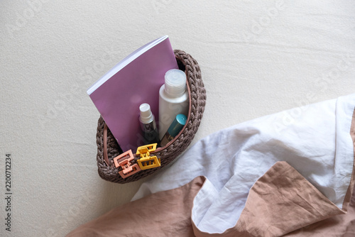 Woven basket containing hair care products, a comb, and a notebook placed on a bed with beige bedding and a light-colored surface.