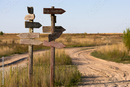 Weathered wooden signposts stand at a fork in a winding dirt road, symbolizing choices and an uncertain path in a natural, open landscape under a clear sky.
