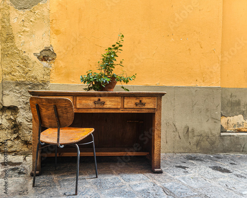 A vintage wooden desk with a potted plant and a wooden chair are set on a stone pavement against a rustic yellow wall in a quiet, narrow outdoor Italian street alley.
