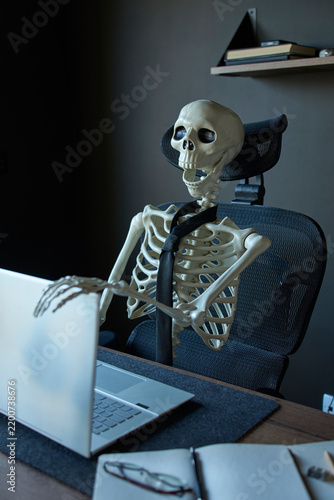Obraz na płótnie Skeleton wearing black tie sitting at office desk with one hand resting on lapto