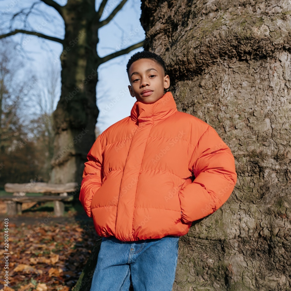 Fototapeta premium Young boy in bright orange puffer jacket leaning on a tree in an autumn park portrait for seasonal banner and background use