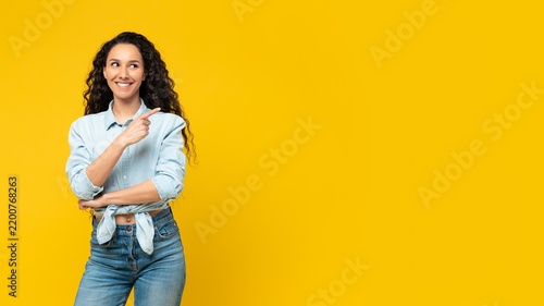 Smiling Young Latina Woman Pointing At Copy Space With Cheerful Expression, Standing In Front Of Yellow Background. Displaying Excitement And Confidence In Business Concept Environment.
