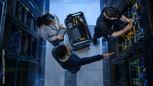 Three IT technicians inspect server racks from a data center aisle, working on network cables and equipment beside a rolling cart, illustrating infrastructure maintenance and network operations