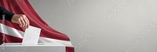 Voter inserts a ballot into a voting box during elections in Latvia. Latvian flag fills the polling backdrop. Democracy and civic participation concept.