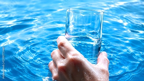 Hand lifting water glass from sparkling blue swimming pool edge
