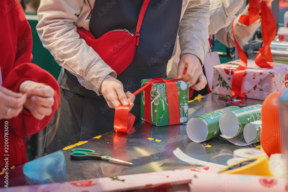 Obraz premium Hands Making Bow on Wrapped Christmas Present. Person finishes wrapping green Christmas box present with wide red ribbon at festive work table. Close-up handmade gift preparation, holiday packaging