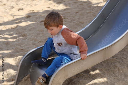 Obraz na plátně Little boy sliding down a playground slide in a sandy park on a sunny day