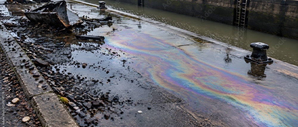 Obraz premium Iridescent diesel film spreading across wet concrete of a canal lock apron beside wreckage in light rain