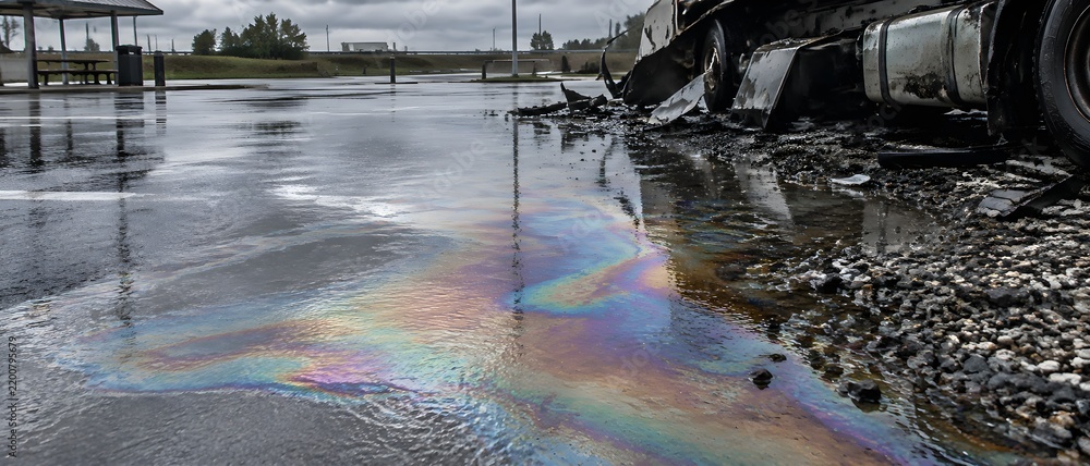 Obraz premium Iridescent diesel film spreading across wet asphalt at a roadside rest area beside wreckage