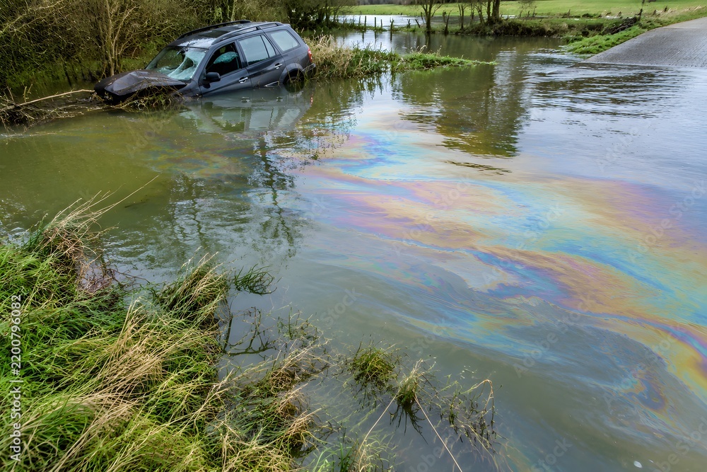 Obraz premium Iridescent diesel film spreading across floodwater in a flooded ford beside a stranded wreck in a river meadow
