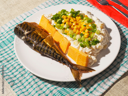 White plate holds a grilled fish  mashed potatoes with corn and green onions  and a side of fried plantain. Bright red napkin with cutlery rests beside the plate on a textured placemat
