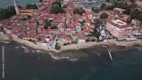 High wide drone shot flying along medieval stone fortification walls, rocky shore with ocean waves, and terracotta rooftops of old town Novigrad Istria