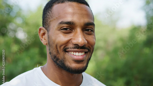Smiling african american man in white t-shirt posing naturally outdoors with green foliage background