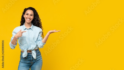 Smiling Young Hispanic Woman Pointing With Hand In Front Of Yellow Background, Showcasing Product Or Service, Energetic And Happy Emotion, Ideal For Advertising Or Marketing Purposes.