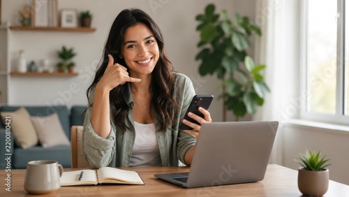 Smiling woman at a home desk makes a phone-call gesture while holding a smartphone, with an open laptop, notebook and coffee mug in a bright, cozy living room workspace