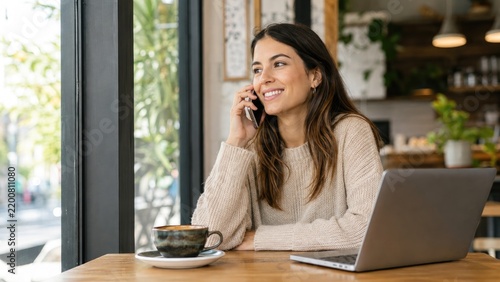 Smiling woman talking on phone in a cozy cafe with laptop, coffee, and natural daylight