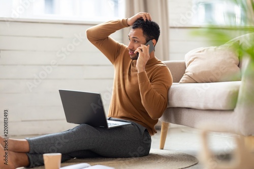 Worried Black Man In His 30s Sitting On Floor, Taking Phone Call While Using Laptop At Home Office, Appearing Stressed And Concerned About Work. Focused On Troubleshooting Business Issues In